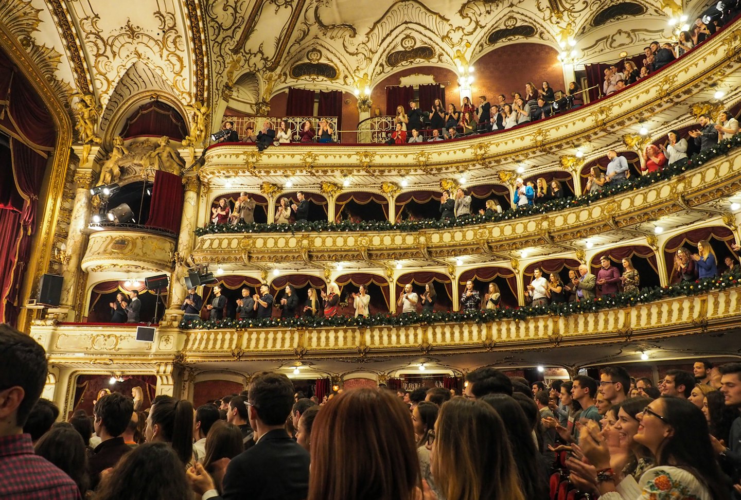 Un pubblico teatrale entusiasta che applaude in ornate balconate a più livelli.