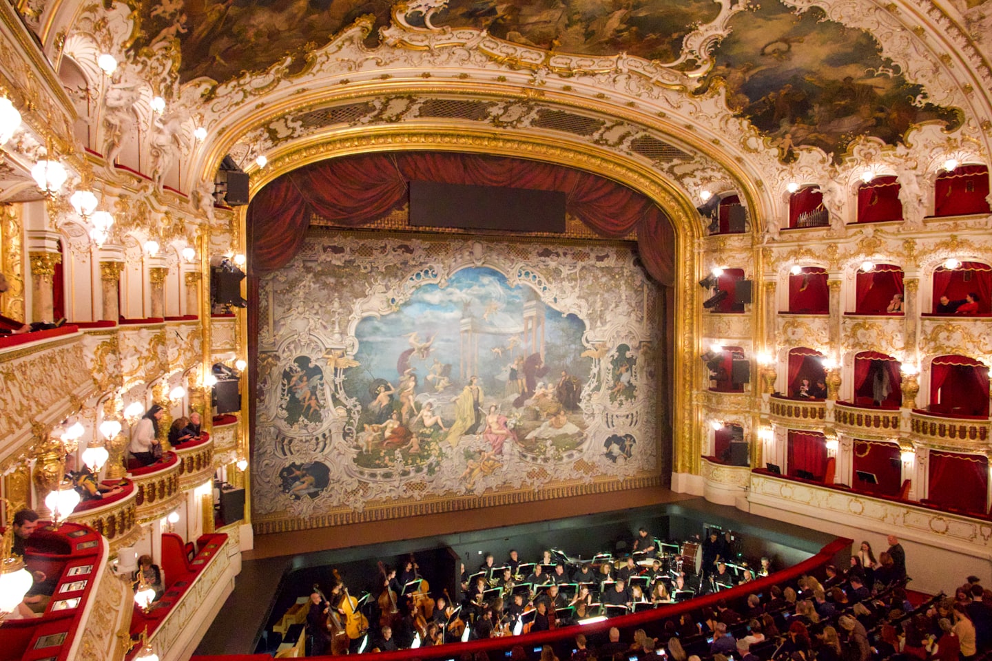 Un'orchestra si esibisce all'interno di uno storico teatro dell'opera ornato e a più livelli.